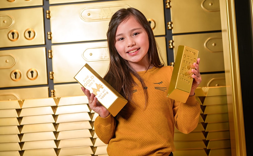 Smiling girl standing in bank vault, holding two gold bullion bars.
