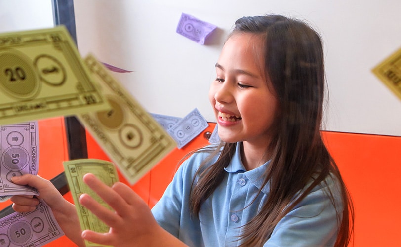 Girl in school uniform in cubicle, smiling and reaching for MONOPLY Money bank notes as they fly about her